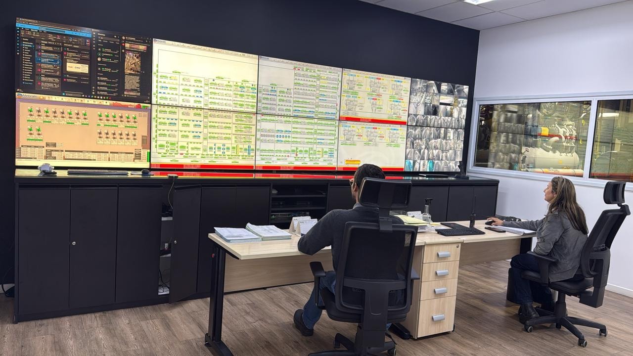 Sala de controle da planta de refrigeração: parede de telas com sinóticos dos sistemas, dois operadores em posto de trabalho e uma janela com vista direta para a casa de máquinas